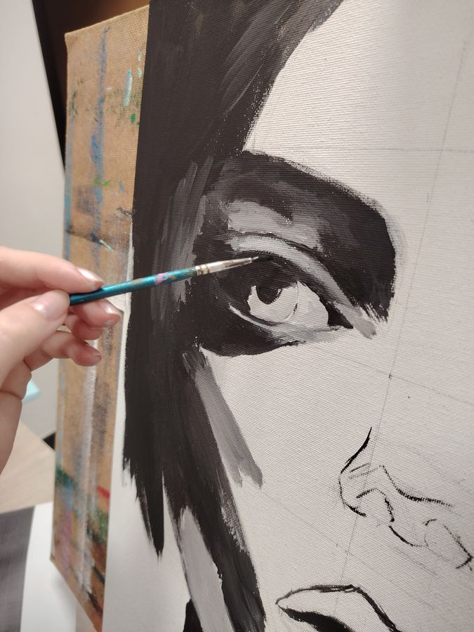 A hand with a fine brush paints the eye of a large black-and-white portrait on canvas.