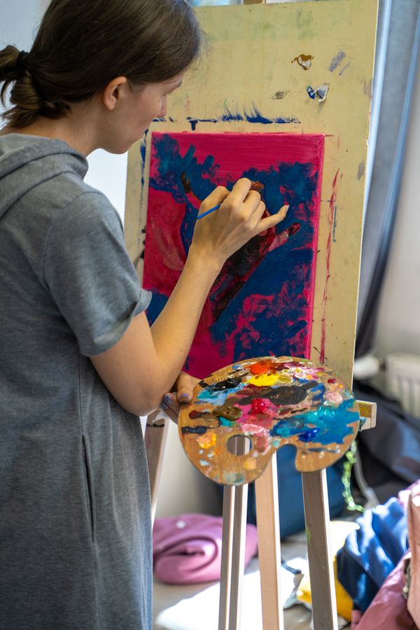 The teacher paints at a wooden easel, holding a palette full of colour.