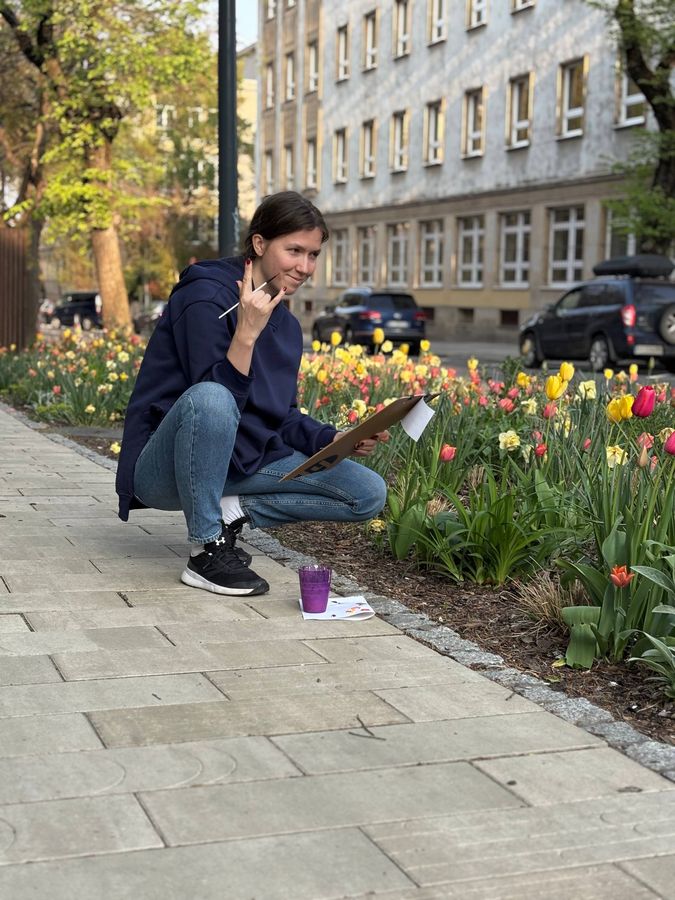 The teacher crouches by a tulip bed in the city, smiling, sketching on a clipboard.