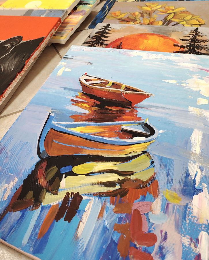 Finished acrylic canvases on the floor, led by a painting of two boats reflected in blue water.