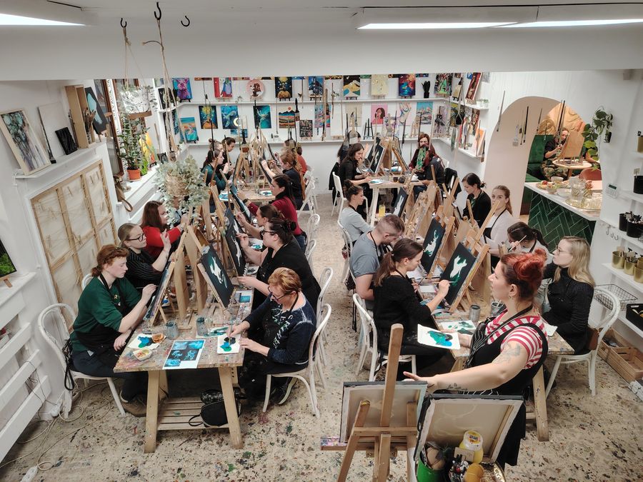 A large group of adults paints at long tables of easels in a bright, gallery-like room.