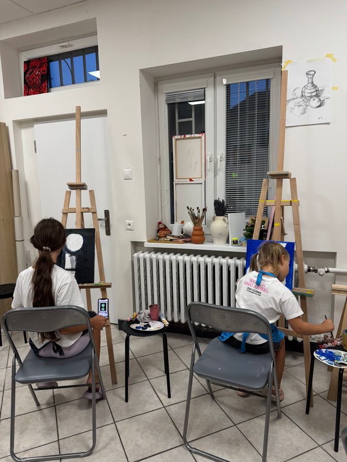 Two girls paint at easels in a bright studio with French windows and still-life drawings on the wall.