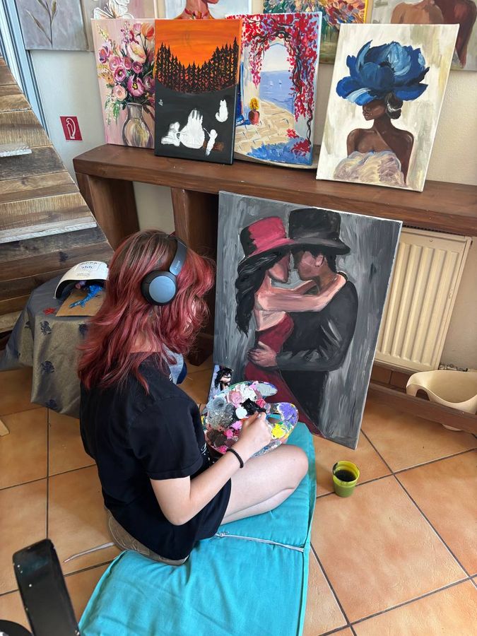 Tínedžerka so slúchadlami sedí na vankúši a maľuje pár v klobúkoch, nad ňou hotové plátna.