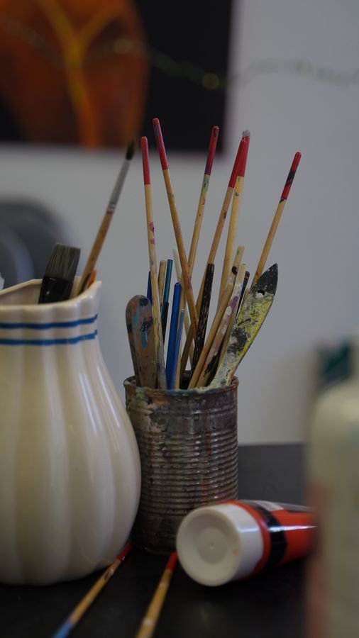 Still life: paint-stained brushes in a tin next to a white vase and a paint tube.