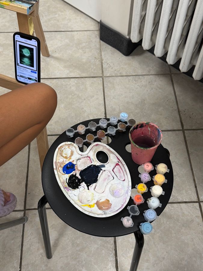 Paleta, kelímky s farbou a pohár s vodou na čiernej stoličke, pohľad zhora.