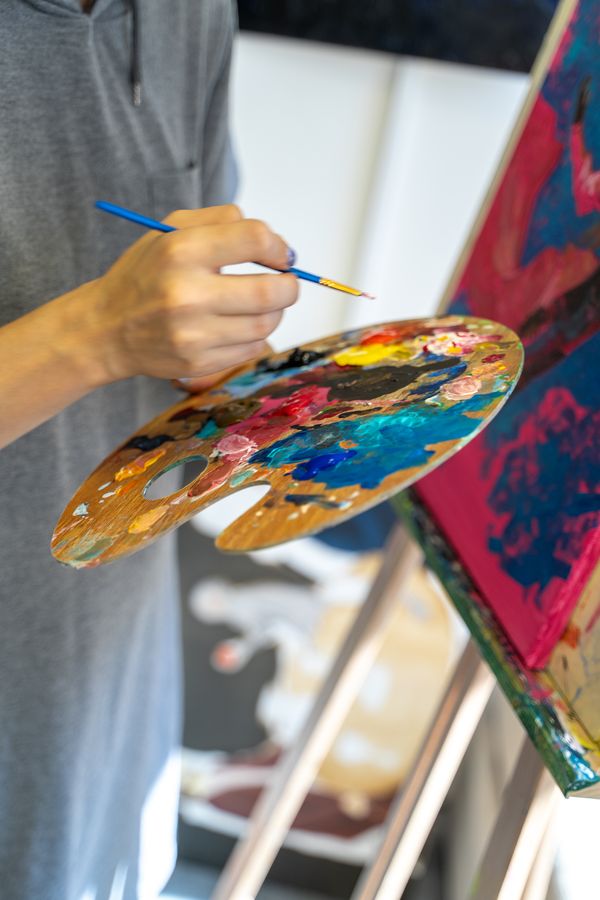 A hand mixes paint on a well-used wooden palette next to the pink canvas.