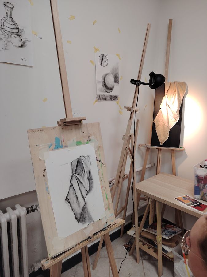 An academic drapery study in graphite on an easel under a clamp lamp, with still-life drawings on the wall.