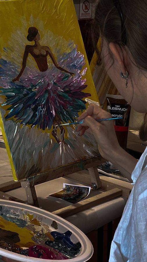 A participant adds palette-knife strokes to a ballerina’s tutu on a bright yellow canvas.