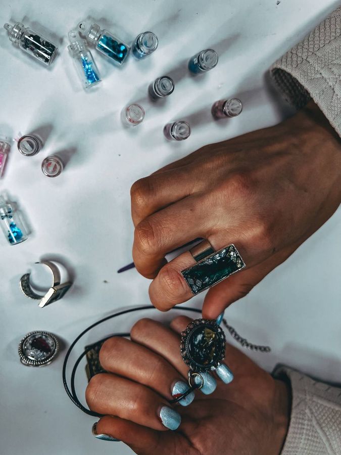 Hands hold freshly made resin rings and a pendant above vials of glitter.