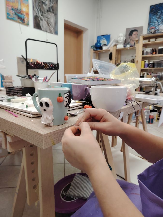 Hands shape a small polymer-clay ghost onto a mug at a studio workbench.