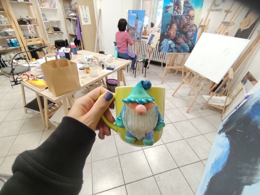 A hand holds up a mug with a polymer-clay gnome in front of a wide view of the studio.