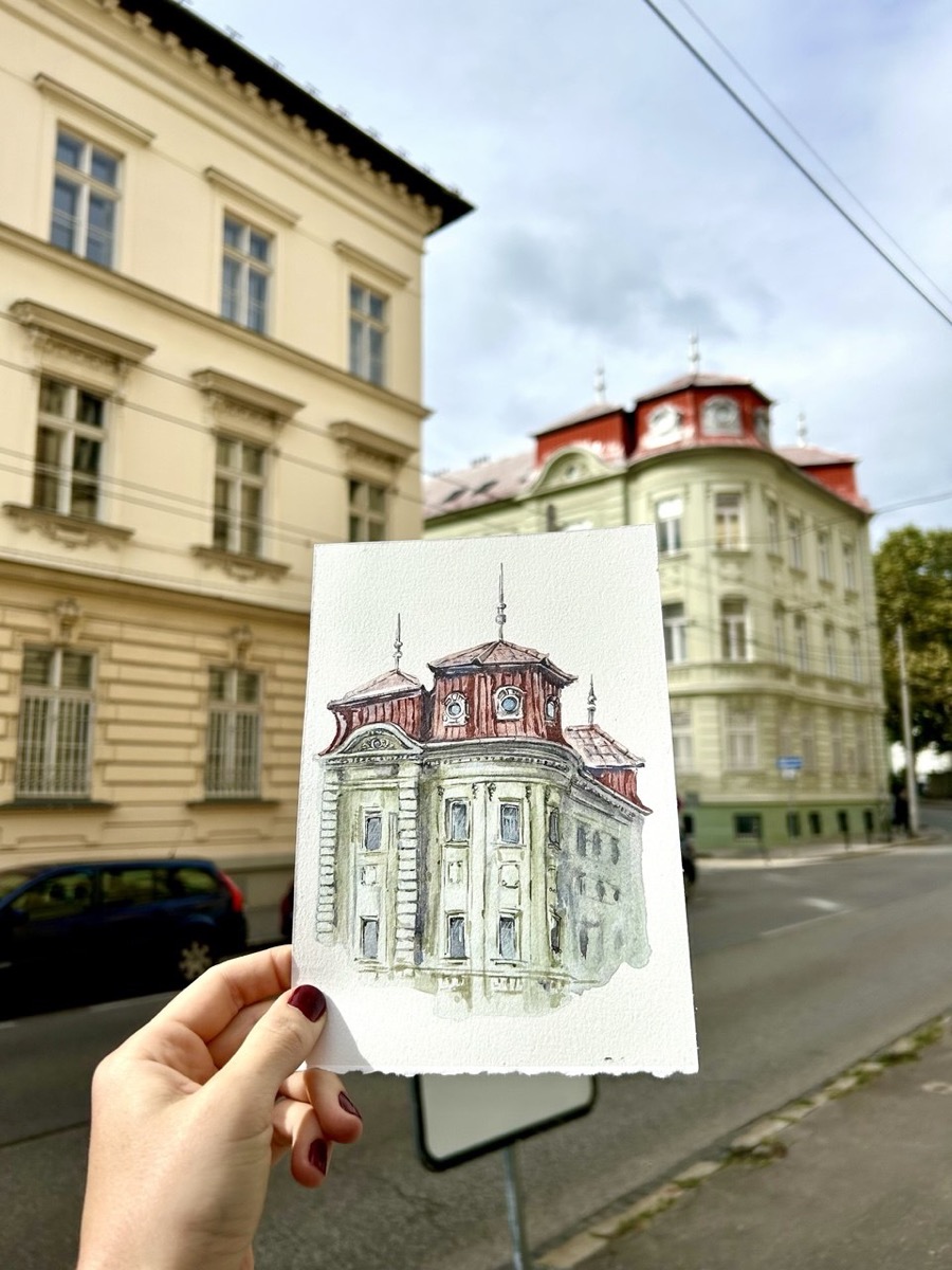 Ruka drží akvarelovú skicu nárožnej budovy s červenými vežičkami priamo pred skutočnou budovou v Bratislave.
