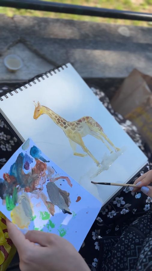 Hands hold a sketchbook with a freshly painted watercolour giraffe and a sheet of colour swatches on a sunny bench.